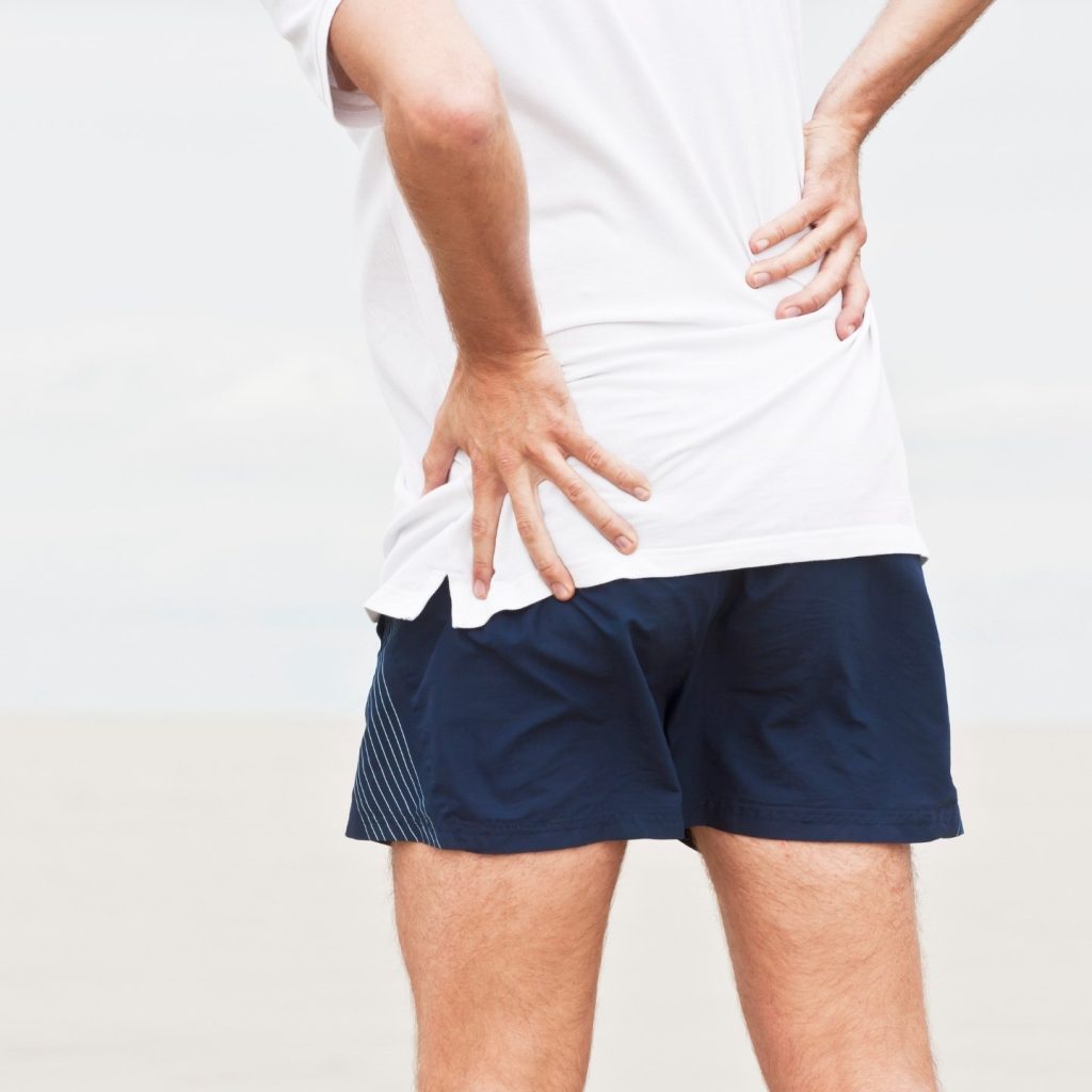 Man in white shirt and blue shorts clutching hip and back because of osteoarthritis and joint pain