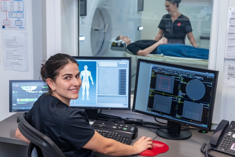 Radiographer sets computer for occupational imaging with radiographer and patient on CT scanner in background.